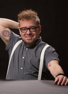 Transgender rocker Lenny Zenith seated with grey shirt and white suspenders